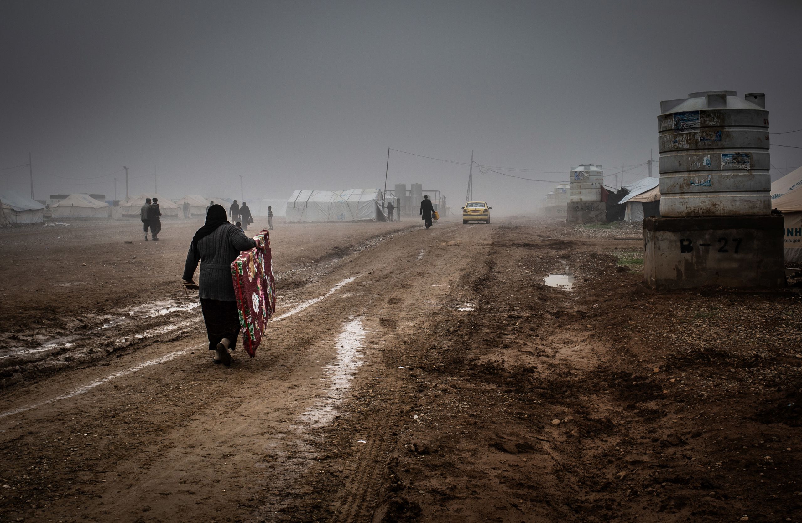 A displaced woman carrying a mattress in the muddy alleys of Hamam al Halil camp, Iraq. (Tom Peyre-Costa /NRC)A displaced woman carrying a mattress in the muddy alleys of Hamam al Halil camp, Iraq. (Tom Peyre-Costa /NRC)
